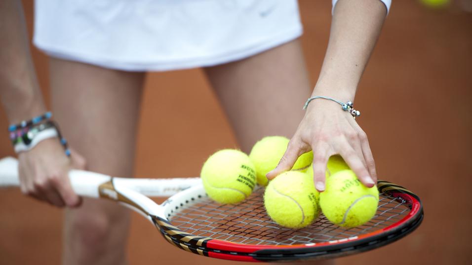 Hand greift mehrere gelbe Tennisbälle auf einem Tennisschläger, während die andere Hand die Schlägerhaltung stabilisiert.